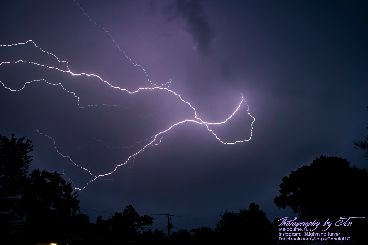Lightning photograph by Jen (@lightninghunter): bright branching lightning arcs sideways across a purple night sky in long, looping channels above silhouetted trees and a utility pole.