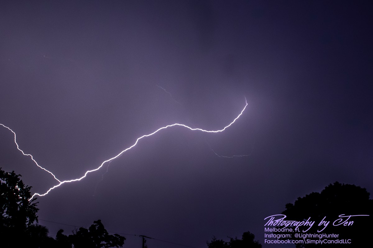 Lightning photograph by Jen (@lightninghunter): a single bright lightning channel snakes in a wide S-curve across a hazy violet sky above dark tree silhouettes.